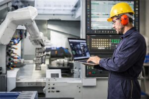 Técnico con casco y protección auditiva usando laptop para controlar un robot industrial en planta de automatización
