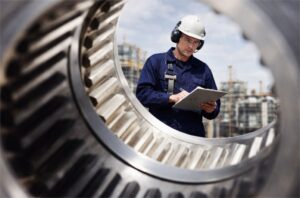 Ingeniero industrial con casco y protección auditiva inspecciona engranajes grandes en planta industrial de automatización