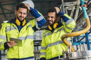 Dos operarios con chaquetas reflectantes y protección auditiva revisan una tableta frente a maquinaria industrial en planta de producción